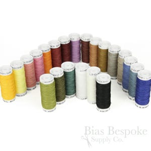 Könnte beinhalten: Eine Sammlung von Leinengarnspulen in verschiedenen Farben, darunter Gelb, Rosa, Grün und Blau. Die Spulen sind in einem Bogen angeordnet, wobei der Markenname "Londonderry Linen" auf einigen sichtbar ist. Der Hintergrund ist weiß.