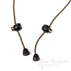 May include: Two brown leather cords with black wooden toggles and brass accents. The cords are braided and have a tassel at the end.