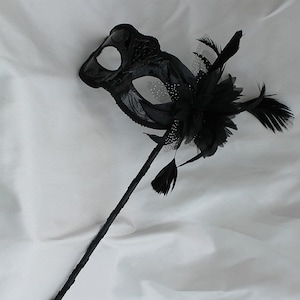 Máscara de baile de máscaras preta deslumbrante em um bastão preto com uma flor feita de pétalas de tecido preto, penas e organza preta e branca