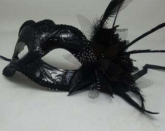 May include: A black masquerade mask with a feathered and floral embellishment. The mask has a delicate design and a glittery finish.