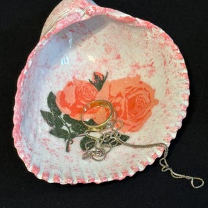 May include: A decorative seashell-shaped trinket dish. The dish is white with pink accents and a floral rose design. A gold ring and silver necklace rest inside the dish. The dish is on a black surface.