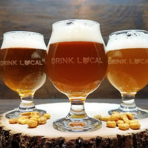 May include: Three clear glass beer goblets with a brown beer inside. Each goblet has the text "DRINK LOCAL" etched on the side with a heart and the outline of the state of Ohio. The goblets are sitting on a wooden slice with a pile of corn kernels on each side.