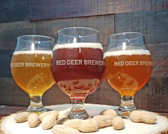 May include: Three glasses of beer from Red Deer Brewery. The glasses are filled with different colored beers: amber, brown, and orange. The glasses are sitting on a wooden surface with peanuts scattered around them.