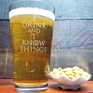 May include: A clear glass beer mug with the text "I drink and I know things" etched on the side. The mug is filled with a golden-colored beer and is sitting on a wooden surface next to a small white bowl filled with cashews.