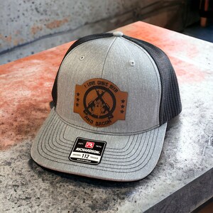 May include: A gray and black trucker hat with a brown leather patch that says "I love guns, beer and bacon". The patch has a crossed rifle design and a pig inside the circle. The hat has a black mesh back and a gray front with a Richardson 112 adjustable label.