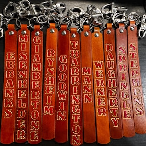 May include: A collection of ten tan leather keychains, each featuring a silver-toned metal swivel snap hook attached by a rivet. Each leather strap is embossed with a unique surname in red lettering, including Eubanks, Misenhelder, Giambertone, Bysheim, Goodwin, Tharrington, Mann, Werner, Daugherty, Springle, and Stephens.