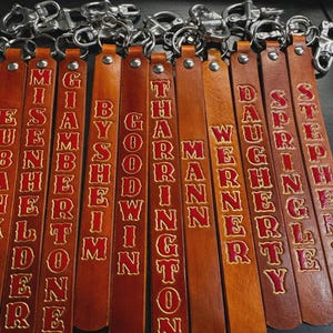 May include: A collection of leather keychains with silver-colored clasps. Each keychain is a different shade of brown and features a name embossed in red lettering. The names include: MISEMB, BYSHERIM, THARMAN, and others.