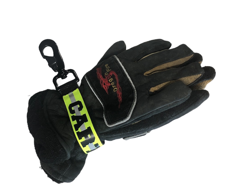 May include: Black firefighter gloves with a yellow and reflective 'CAPF' strap and a black carabiner clip. The gloves have a brown leather palm and a red and white 'Dragon Fire' logo.