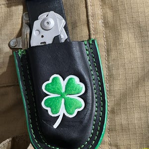 May include: Black leather multi-tool sheath featuring a green and white four-leaf clover design. The sheath has green stitching and is attached to a black belt. A silver multi-tool is partially visible.