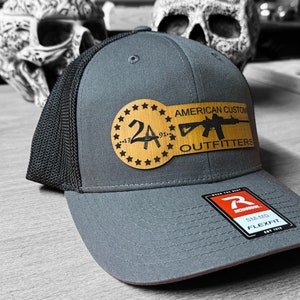May include: A grey and black mesh trucker hat with a gold patch featuring a stylised rifle and the text "2A", "AMERICAN CUSTOM OUTFITTERS", and a circle of stars. The hat has a red label with the text "Richardson" and "Flexfit" on the underside of the brim.