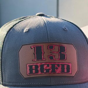 Könnte beinhalten: Eine graue und schwarze Mesh-Trucker-Kappe mit einem braunen Leder-Patch, auf dem "13 BCFD" in schwarzen und roten Buchstaben steht.