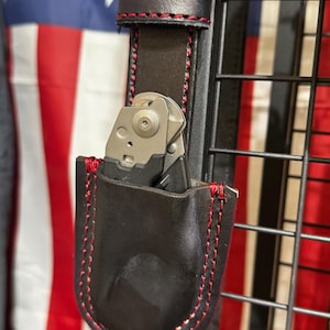 May include: A dark brown leather sheath with vibrant red stitching, designed to hold a folding tool. The sheath is attached to a matching leather belt loop, also featuring red stitching, and contains a metallic folding tool with a silver finish.