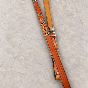 May include: A tan leather strap featuring the name R. SMITH embossed in black lettering. The strap includes silver-toned metal hardware, including a buckle and rivets, with a smooth finished front and a textured, natural suede-like underside.