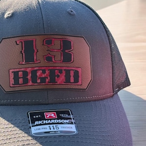 May include: A gray and black trucker hat with a brown patch that says "13 BGFD" in red and black lettering. The hat has a black mesh back and a Richardson logo on the front.