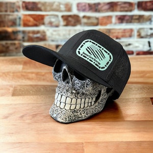Puede incluir: Gorra de camionero negra con parte trasera de malla y visera plana. La gorra presenta un parche rectangular con un diseño de bandera estadounidense verde menta y el texto "American Custom Outfitters". La gorra está sobre una calavera decorativa.