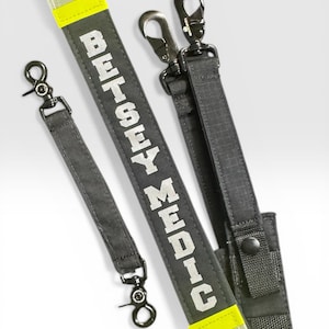 May include: A dark gray tactical strap set featuring the text BETSEY MEDIC in white block letters. The straps include bright neon yellow reflective strips at the ends and black metal swivel snap hooks for attachment, with a snap button closure.