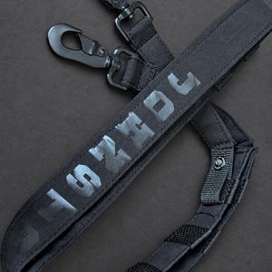 May include: A black camera strap featuring the text "SHOGUN" in a reflective gray font. The strap includes two black metal swivel snap hooks attached to nylon webbing, with adjustable sections secured by hook-and-loop fasteners for length control.