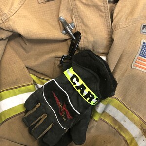 May include: A black leather fire glove with a yellow reflective strap that says "CARI" on it. The glove is attached to a silver carabiner clip and is hanging on a brown fire jacket with a yellow and white stripe.