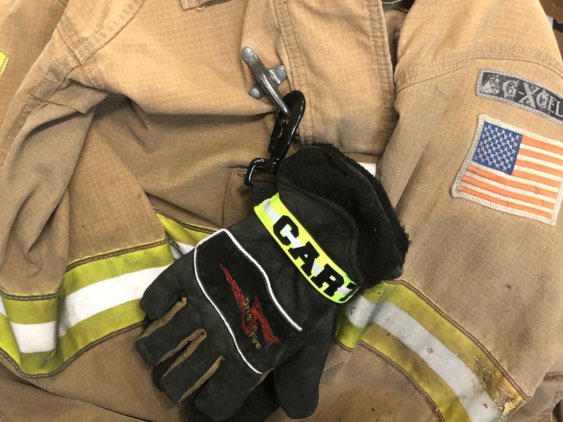 May include: A black leather fire glove with a yellow reflective strap that says "CARI" on it. The glove is attached to a silver carabiner clip and is hanging on a brown fire jacket with a yellow and white stripe.