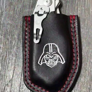 May include: Black leather multi-tool sheath with red stitching and a white Darth Vader graphic. A silver multi-tool is partially visible. The sheath is designed to hold the multi-tool.