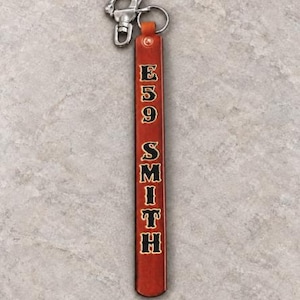 May include: A long, rectangular, reddish-brown leather keychain strap featuring the text E59 SMITH embossed in black with a gold outline. The strap is attached to a silver-toned metal swivel lobster clasp and a circular split ring at the top.
