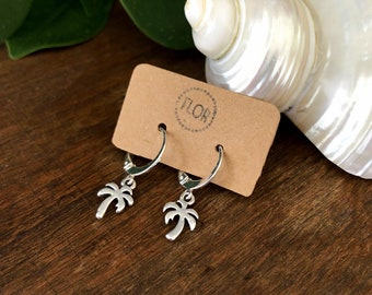 May include: A pair of silver hoop earrings with palm tree charms hanging from them. The earrings are displayed on a brown card with the word "FLOR" printed on it.