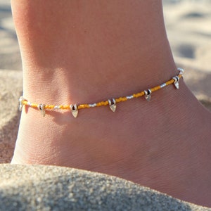 Könnte beinhalten: Fußkettchen mit kleinen, gelben Perlen, silberfarbenen Perlen und dekorativen Anhängern. Der Schmuck wird am Knöchel getragen, mit Sand im Hintergrund.