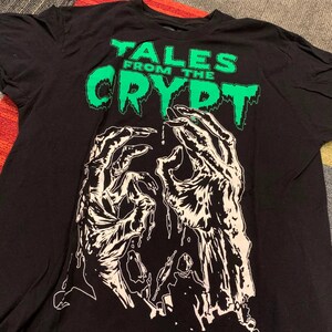 May include: A black short-sleeve t-shirt featuring the green text "TALES FROM THE CRYPT" at the top. Below the text is a large, detailed white graphic illustration of two skeletal, dripping hands reaching upward against the black fabric background.