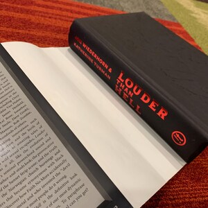 Puede incluir: Un libro de tapa dura negro titulado "Louder Than Hell" de John Wiederhorn y Katherine Turman, con letras rojas. El libro est&aacute; abierto, revelando p&aacute;ginas blancas.
