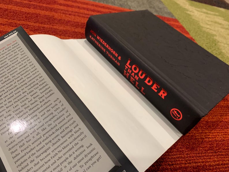 Puede incluir: Un libro de tapa dura negro titulado "Louder Than Hell" de John Wiederhorn y Katherine Turman, con letras rojas. El libro est&aacute; abierto, revelando p&aacute;ginas blancas.