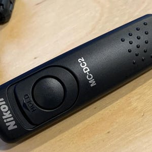 May include: Black Nikon MC-DC2 remote shutter release with a large round button and the word "HOLD" printed on the side. The remote has a textured surface and a series of small holes near the top. A black cable extends from the bottom.