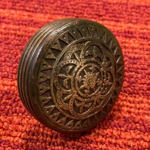 May include: A round, metallic, bronze-toned door knob featuring an intricate, embossed floral pattern in the center, surrounded by a border of triangular shapes and a series of concentric, grooved lines along the outer edge.