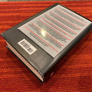 Puede incluir: Un libro de tapa dura con una cubierta negra y texto blanco en la parte posterior. El libro est&aacute; sobre una superficie a rayas rojas y naranjas. El t&iacute;tulo y el autor del libro son visibles.