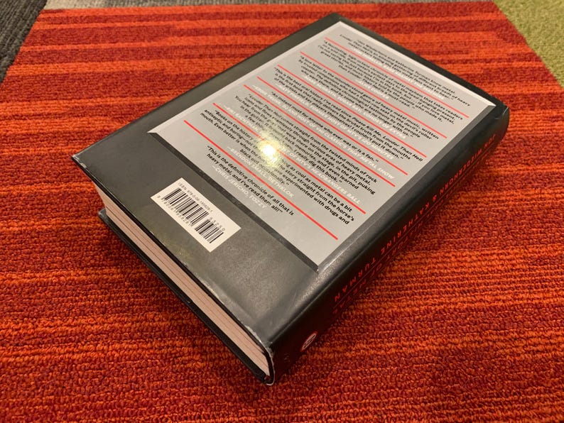 Puede incluir: Un libro de tapa dura con una cubierta negra y texto blanco en la parte posterior. El libro est&aacute; sobre una superficie a rayas rojas y naranjas. El t&iacute;tulo y el autor del libro son visibles.