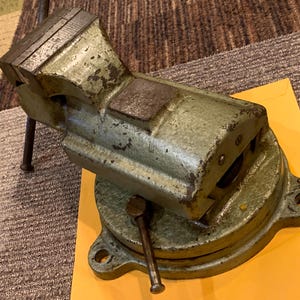 May include: A heavy-duty metal bench vise with a weathered, sage green finish. The tool features a rotating circular base with three mounting holes, a sliding jaw mechanism, and a steel handle used for tightening the grip on workpieces.