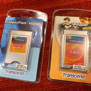 May include: Two Transcend CompactFlash and 5-in-1 adapters in clear plastic packaging. The CompactFlash adapter is on a blue background, while the 5-in-1 adapter is on an orange background. Both have the Transcend logo.
