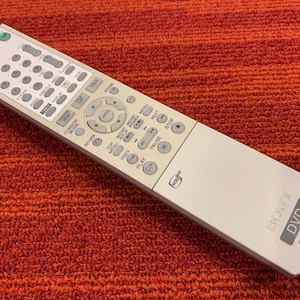 May include: A white Sony DVD remote control with numerous buttons for various functions. The remote features a circular navigation pad and buttons labeled "Play," "Pause," and "Stop." The remote is on a red-orange textured surface.