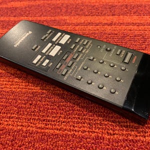 May include: Black Panasonic VCR remote control with numerous buttons for various functions. The remote features buttons for power, channel selection, play, pause, and more. The remote is on a red-orange textured surface.