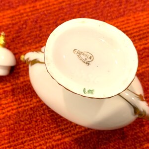 K&ouml;nnte beinhalten: Wei&szlig;er Keramik-Sahneaufgie&szlig;er mit Goldrand und passendem Deckel. Der Sahneaufgie&szlig;er hat eine geschwungene Form und einen Griff. Auf der Unterseite des Sahneaufgie&szlig;ers steht "ERPHILA GERMANY". Der Deckel hat eine goldene Spitze.