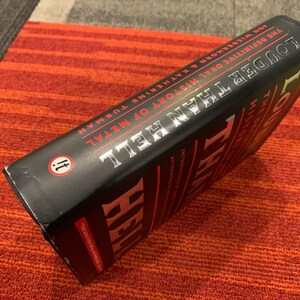 Puede incluir: Un libro negro titulado "Louder Than Hell: The Definitive Oral History of Metal" de Jon Wiederhorn y Katherine Turman. El libro est&aacute; sobre una superficie a rayas rojas y naranjas.