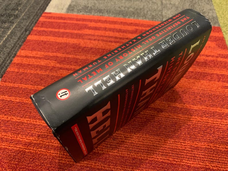 Puede incluir: Un libro negro titulado "Louder Than Hell: The Definitive Oral History of Metal" de Jon Wiederhorn y Katherine Turman. El libro est&aacute; sobre una superficie a rayas rojas y naranjas.