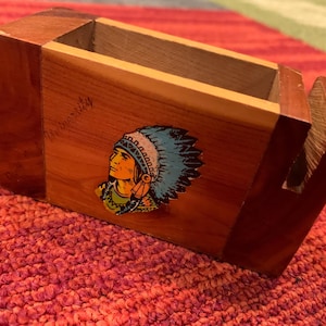 May include: Wooden desk organizer with a Native American chief illustration. The organizer is made of reddish-brown wood and features a compartment and a slot. The illustration is in blue, yellow, and green.