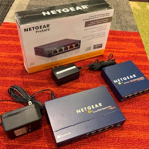 May include: A Netgear ProSafe 5-port Fast Ethernet switch in its original white and orange box, alongside a blue Netgear Fast Ethernet switch, a blue Netgear Dual Speed Hub, and two black power adapters with attached cords and wall plugs.