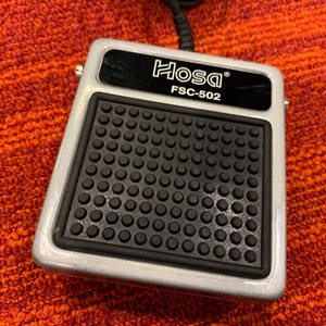 May include: A silver Hosa FSC-502 foot switch pedal with a black rubber pad featuring a grid of small, circular bumps. The pedal has a black label with the Hosa logo and model number. A black cable extends from the top.
