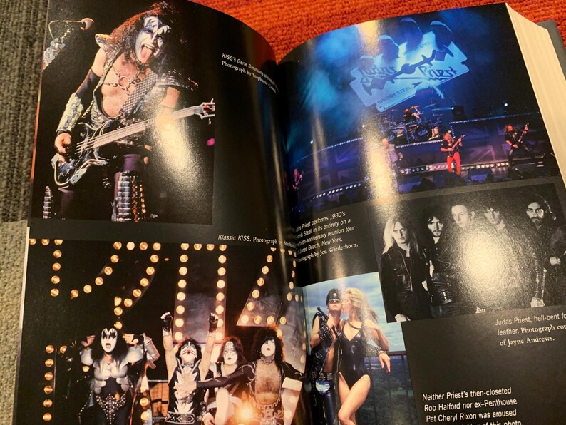 Puede incluir: Una revista con fotos de conciertos de las bandas de rock KISS y Judas Priest. La foto de KISS muestra a un miembro de la banda tocando el bajo. La foto de Judas Priest muestra a la banda actuando en el escenario. La revista est&aacute; abierta sobre una superficie naranja.