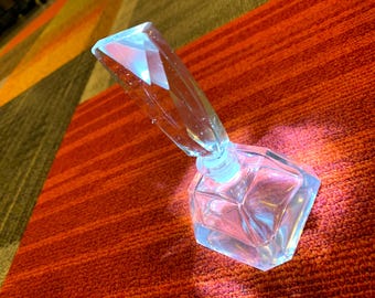 Könnte beinhalten: Ein durchsichtiger Glasparfümflakon mit facettiertem, geometrischem Boden und einem hohen, kantigen, kristallartigen Verschluss. Das Glas reflektiert das Licht und erzeugt helle Glanzlichter sowie einen bläulichen Schimmer am Flaschenhals.