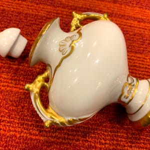 K&ouml;nnte beinhalten: Eine wei&szlig;e Porzellanvase mit goldenen Akzenten, einschlie&szlig;lich Griffen und Zierleisten. Die Vase hat einen Deckel und steht vor einem rot-orangen strukturierten Hintergrund. Die Vase dient wahrscheinlich dekorativen Zwecken.