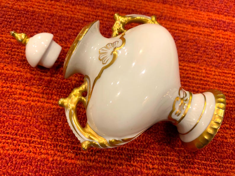 K&ouml;nnte beinhalten: Eine wei&szlig;e Porzellanvase mit goldenen Akzenten, einschlie&szlig;lich Griffen und Zierleisten. Die Vase hat einen Deckel und steht vor einem rot-orangen strukturierten Hintergrund. Die Vase dient wahrscheinlich dekorativen Zwecken.