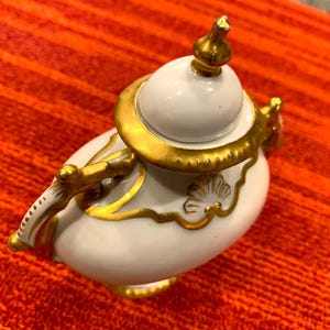 K&ouml;nnte beinhalten: Eine wei&szlig;e Porzellan-Teekanne mit goldenen Akzenten. Die Teekanne hat einen runden K&ouml;rper, einen Deckel mit Knauf und einen gebogenen Griff. Goldene Verzierungen umranden Deckel, Griff und dekorative Elemente. Die Teekanne steht vor rotem Hintergrund.