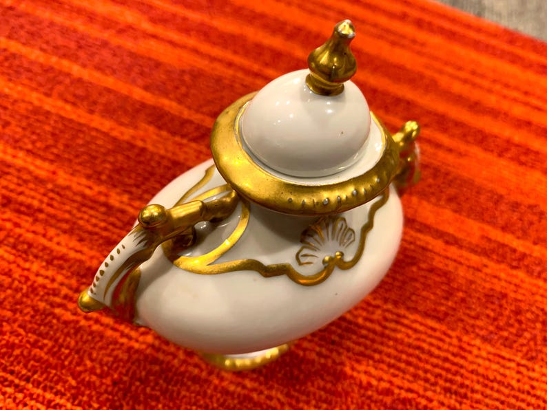 K&ouml;nnte beinhalten: Eine wei&szlig;e Porzellan-Teekanne mit goldenen Akzenten. Die Teekanne hat einen runden K&ouml;rper, einen Deckel mit Knauf und einen gebogenen Griff. Goldene Verzierungen umranden Deckel, Griff und dekorative Elemente. Die Teekanne steht vor rotem Hintergrund.
